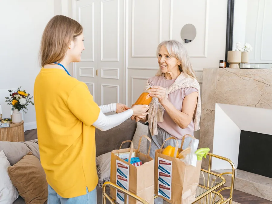 A caregiver handing groceries to an older woman in her living room