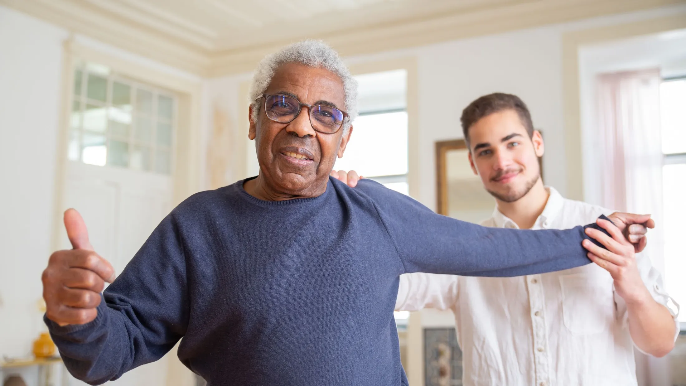 A caregiver steadying a smiling older man as he stretches out his arms at home