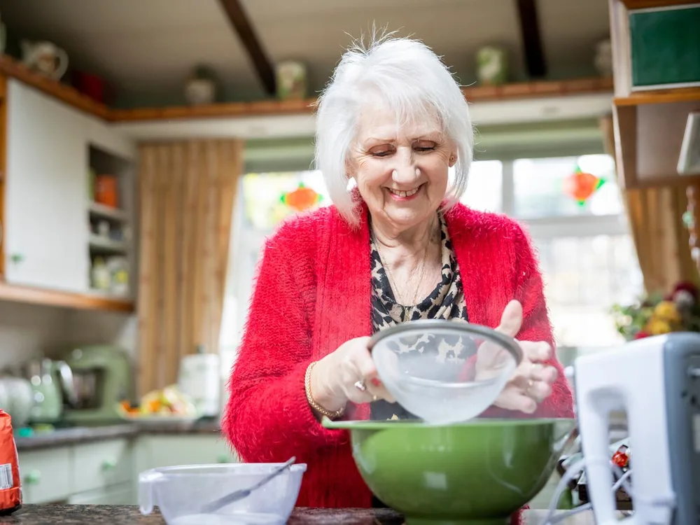 An older woman smiling as she sifts flour while baking in her own kitchen