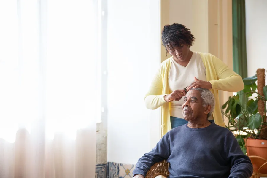 A caregiver gently combing an older man's hair beside a sunlit window
